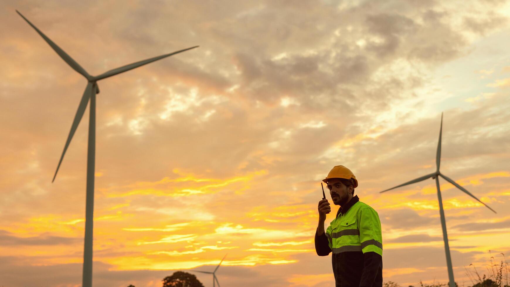 man in a yellow and black safety vest is standing in front of three wind turbines. The sky is orange and the man is holding a walkie talkie photo