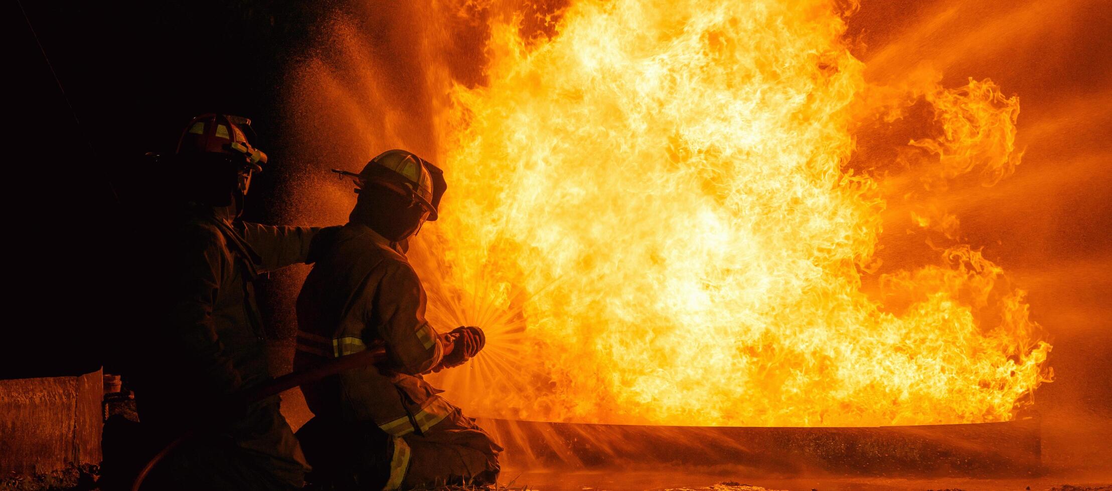 Two firefighters are standing in front of a fire, one of them is holding a hose photo