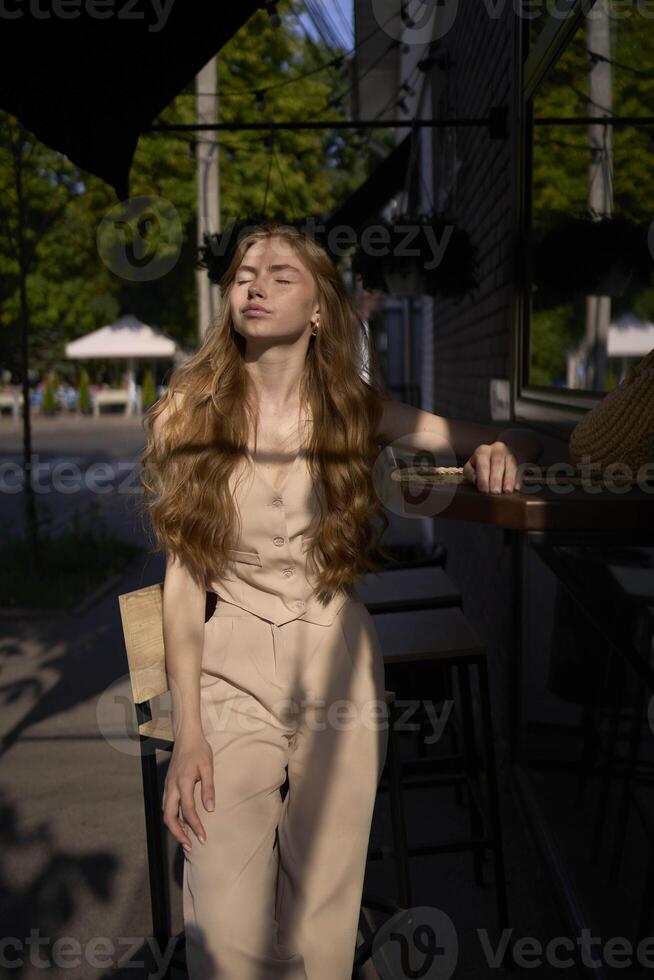 a beautiful teenage girl in a peach fuzz suit with a waistcoat in the old money style sits at the bar outside photo