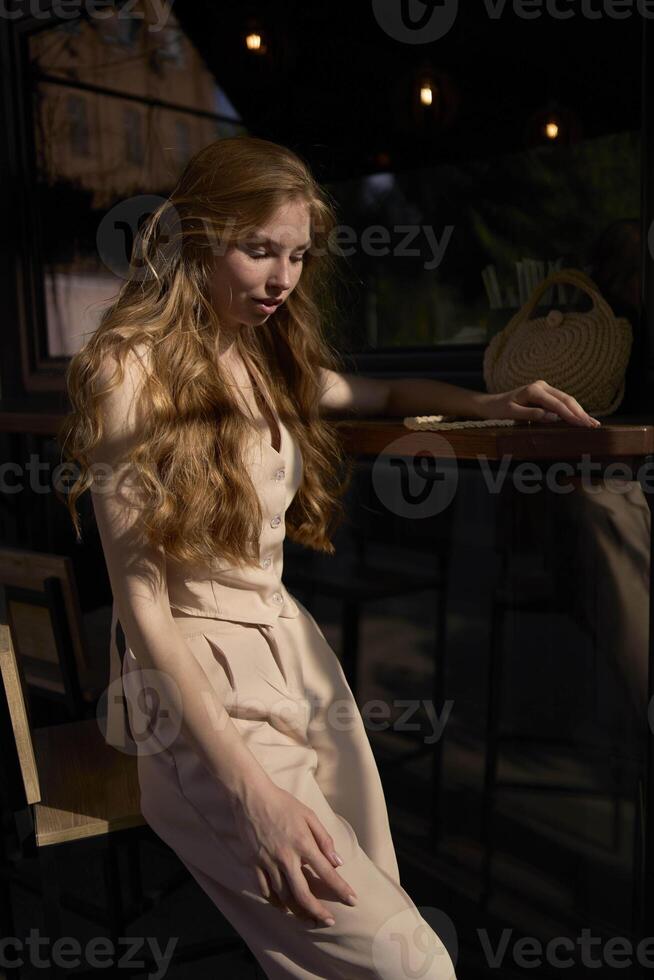 a beautiful teenage girl in a peach fuzz suit with a waistcoat in the old money style sits at the bar outside photo