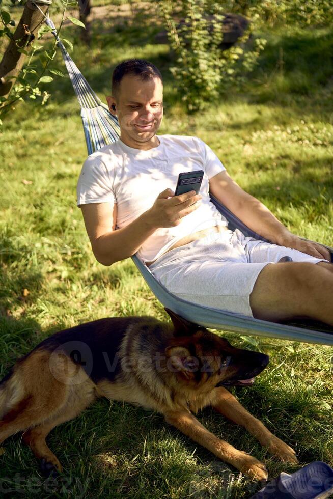 a man rests and listens to music in a hammock, his dog rests nearby photo