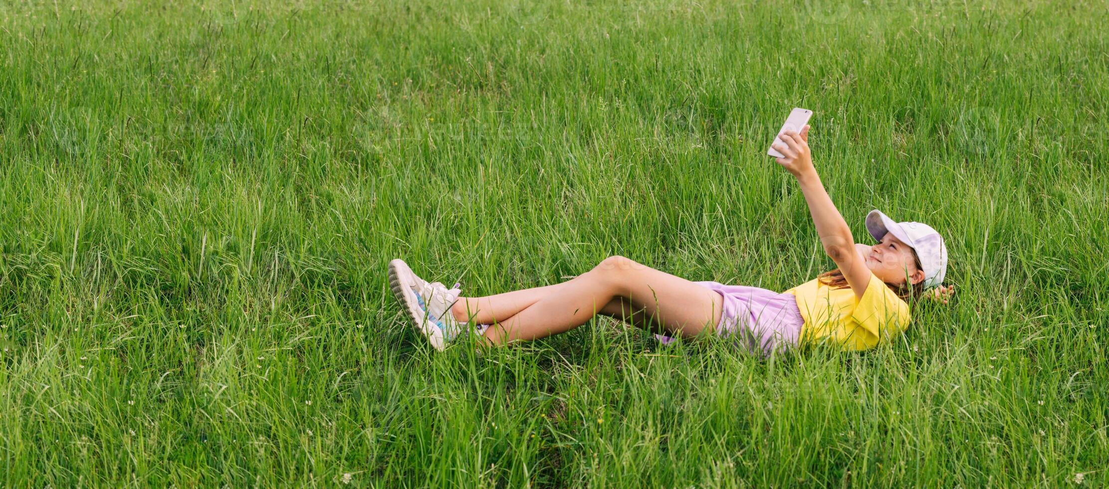 A preteen girl lies on a green grass lawn and takes a selfie on a mobile phone. Horizontal frame photo