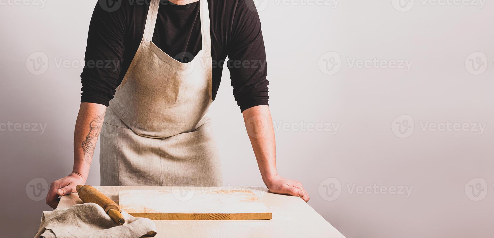Men cook in a cotton apron holds a wooden rolling pin at the kitchen table with a cutting board. Rustic style. photo
