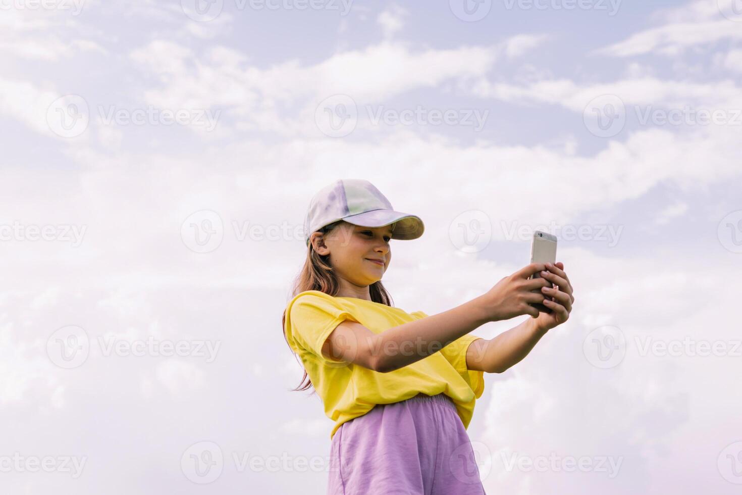 A preteen girl takes a selfie on a mobile phone against a blue sky. Horizontal frame, copy space photo