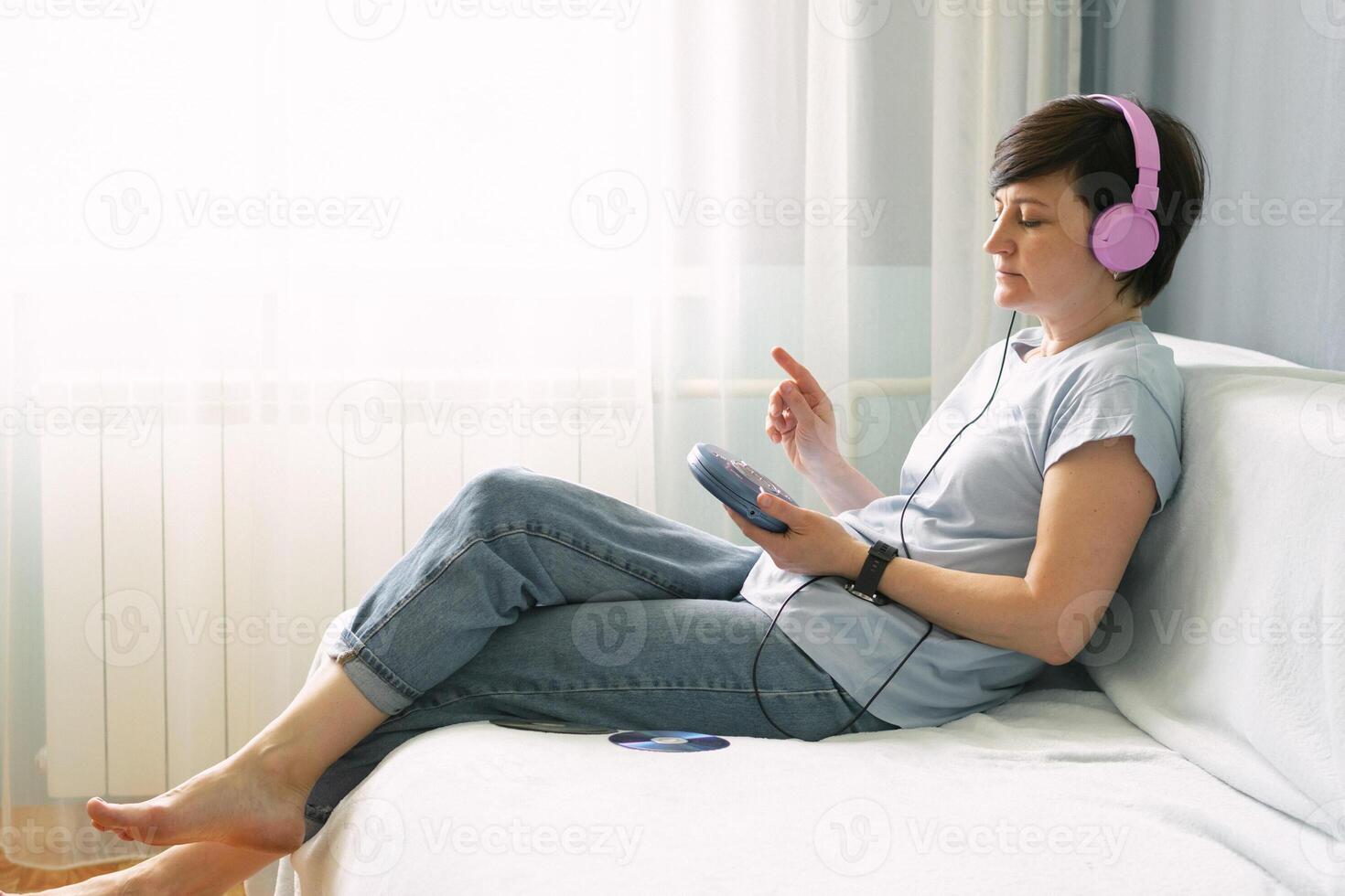 An adult woman in pink headphones sits on the sofa at home and listens to music on the CD players photo