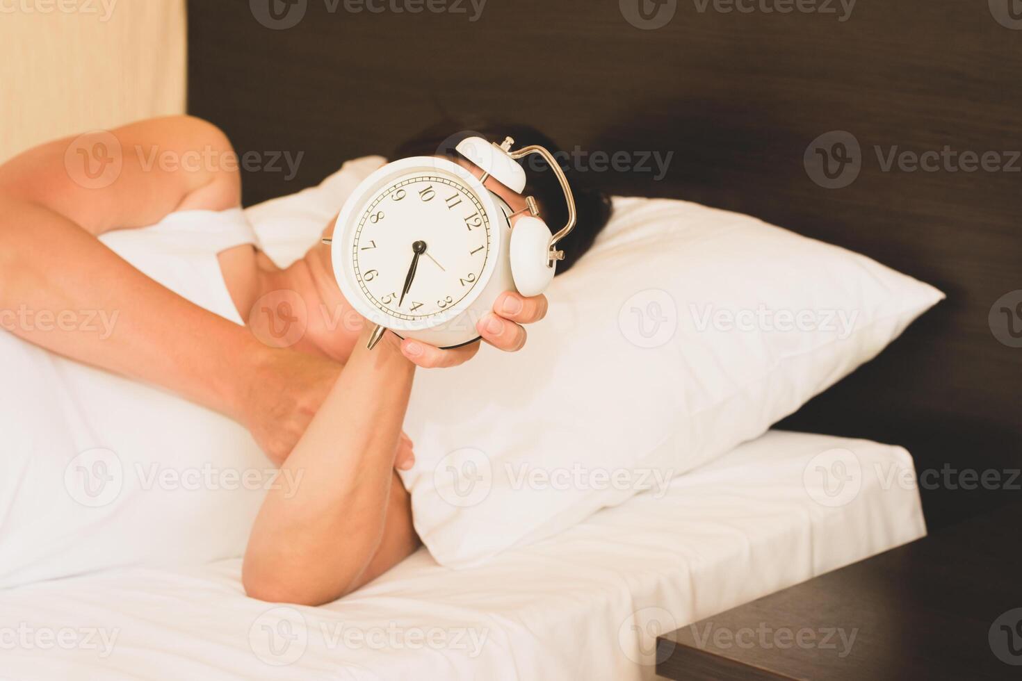 A woman lies in bed with an alarm clock in her hands. Selective focus with focus on the clock. Morning awakening, photo