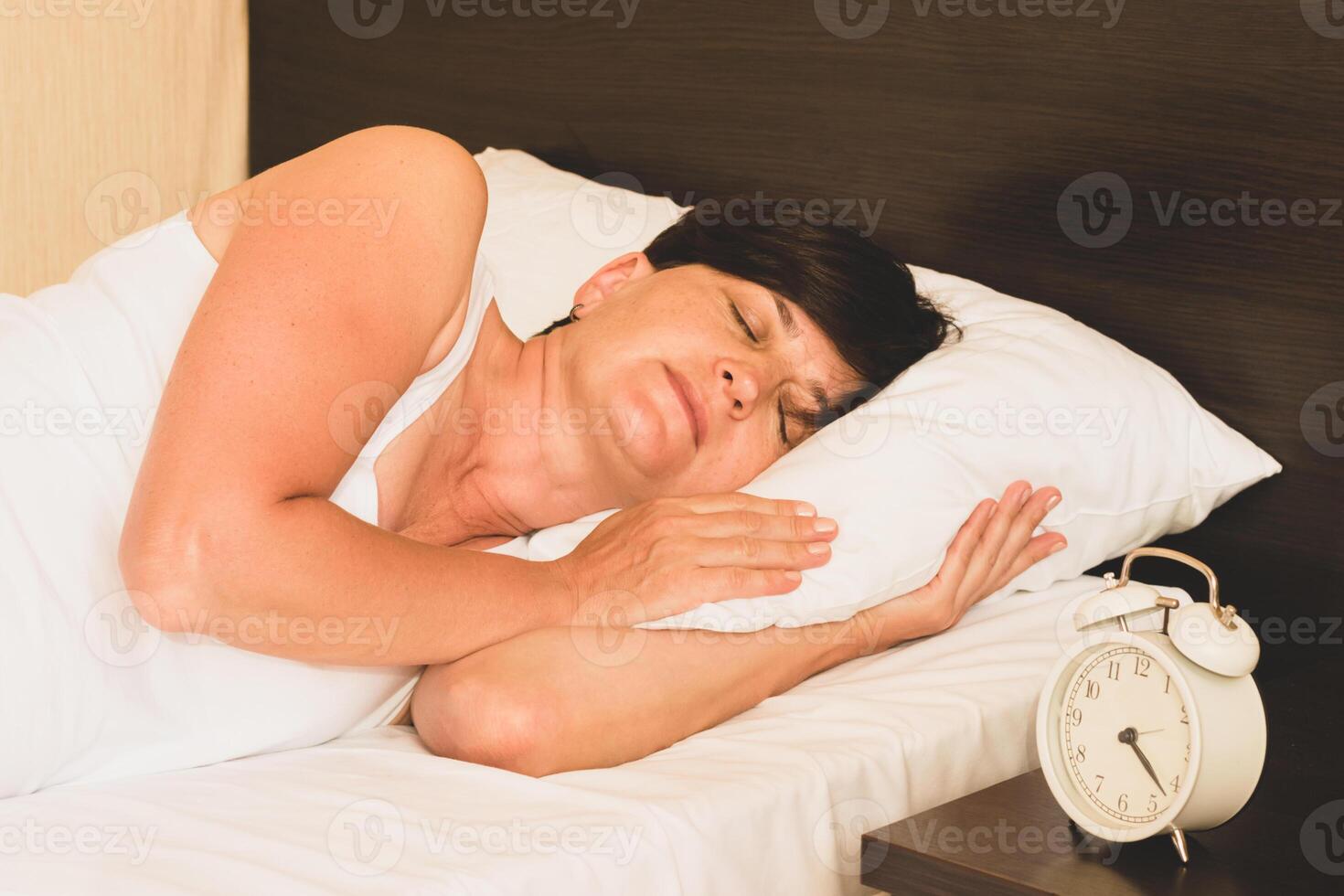Middle-aged woman asleep in bed, alarm clock on the bedside table. Morning awakening, sleep and rest. Women's harmony photo