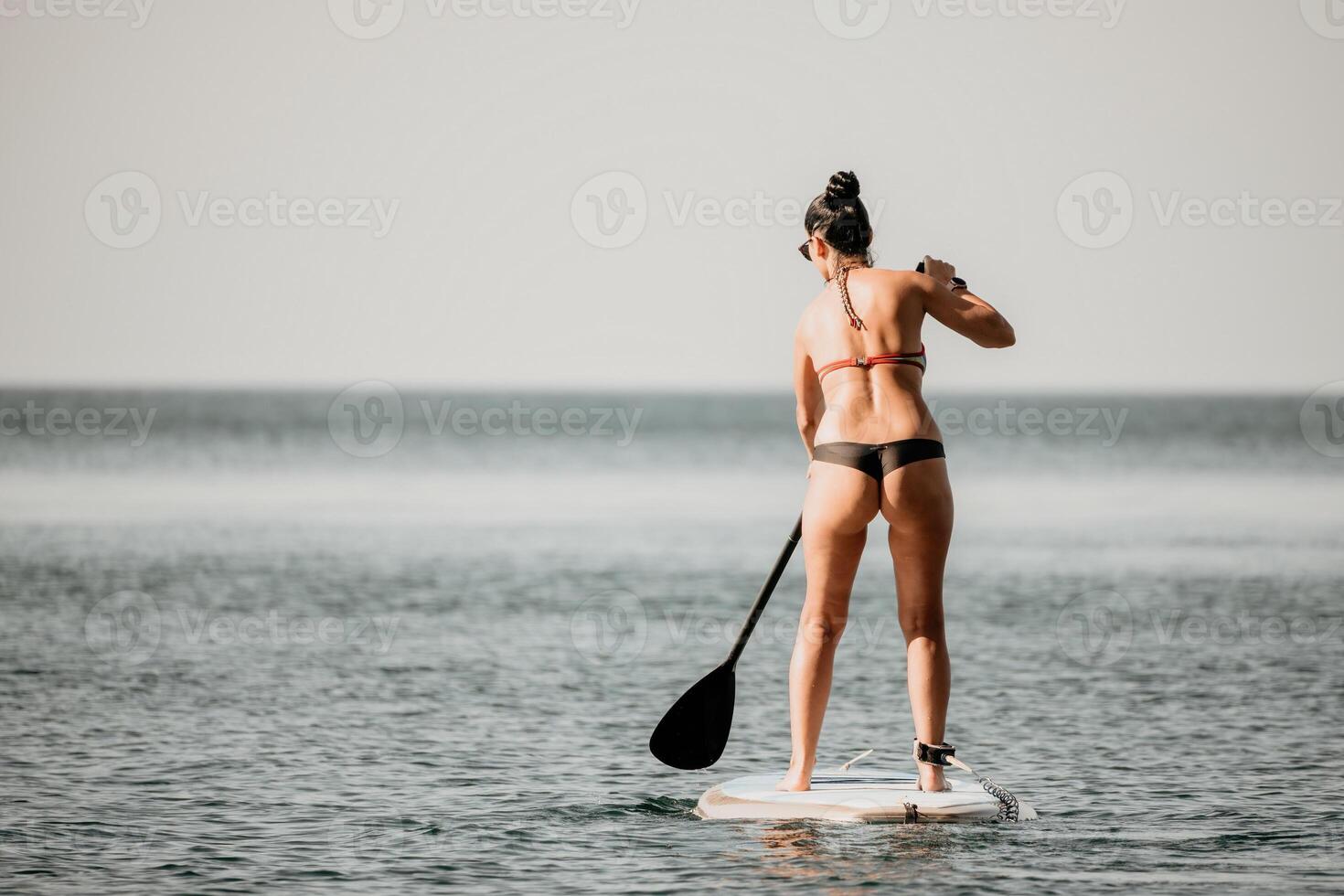 Sea woman sup. Silhouette of happy middle aged tanned woman in rainbow bikini, surfing on SUP board, confident paddling through water surface. Idyllic sunset. Active lifestyle at sea or river. photo