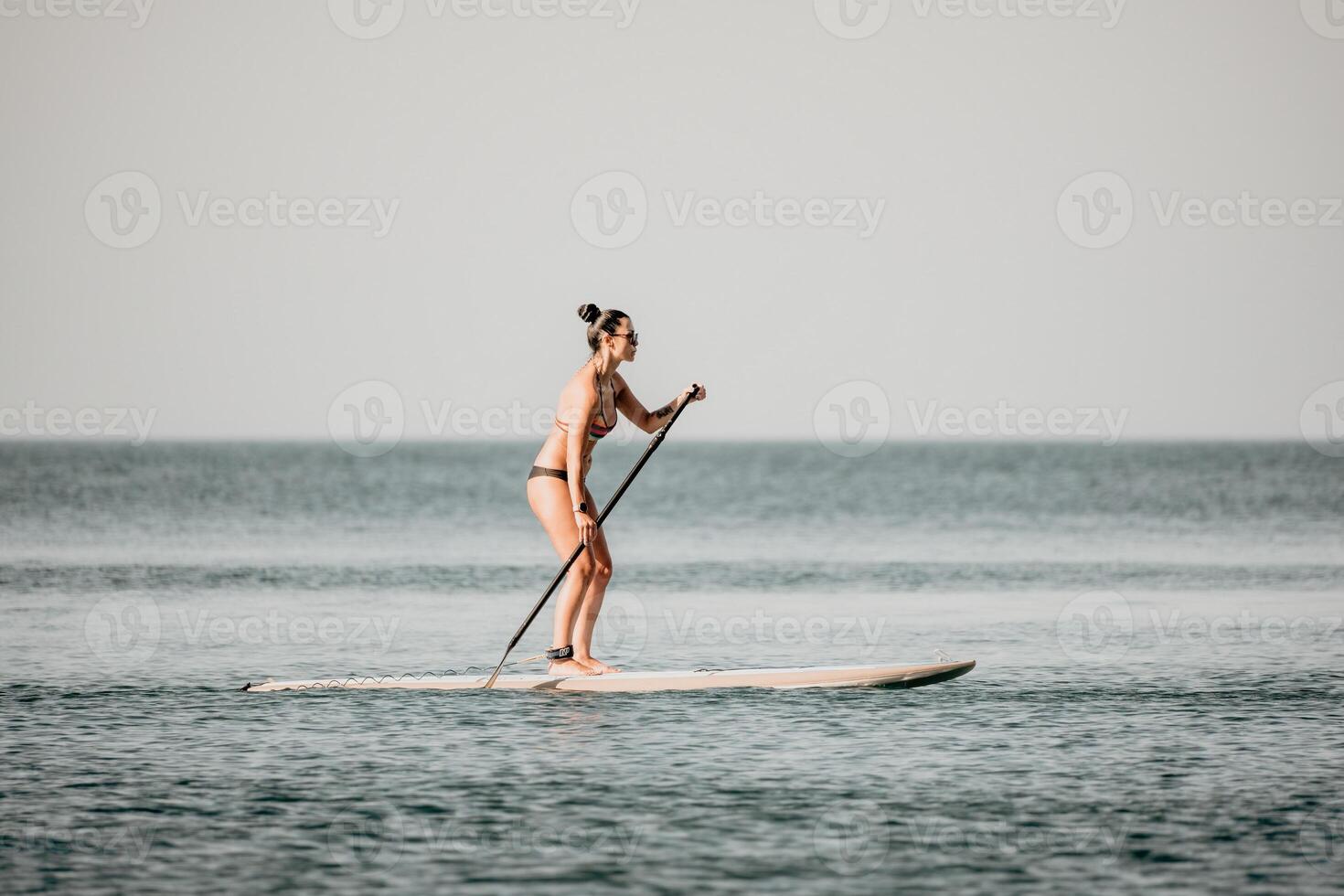 Sea woman sup. Silhouette of happy middle aged tanned woman in rainbow bikini, surfing on SUP board, confident paddling through water surface. Idyllic sunset. Active lifestyle at sea or river. photo