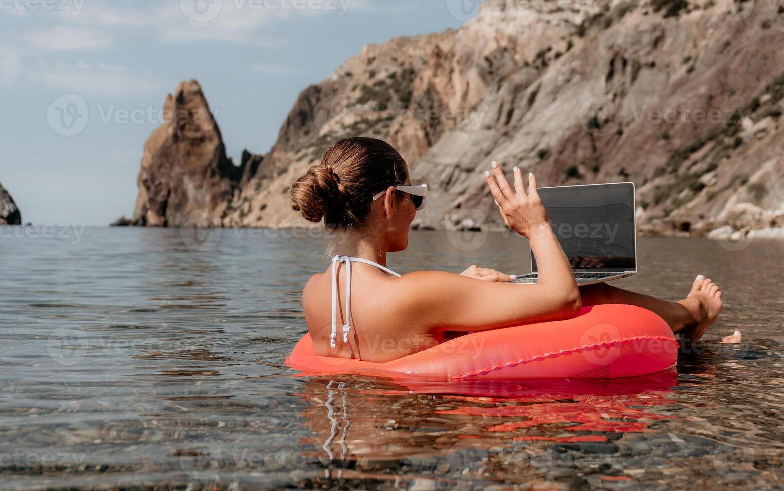 Woman freelancer works on laptop swimming in sea on pink inflatable ring. Happy tourist in sunglasses floating on inflatable donut and working on laptop computer in calm ocean. Remote working anywhere photo