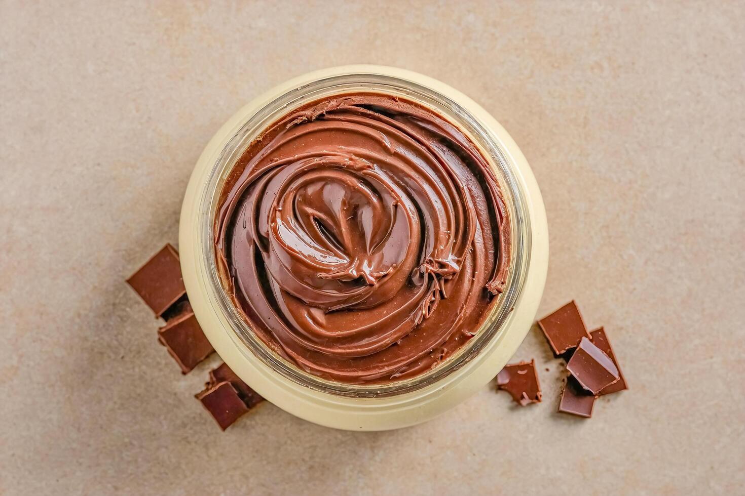 Chocolate spread in a glass jar with chocolate pieces on a beige background photo