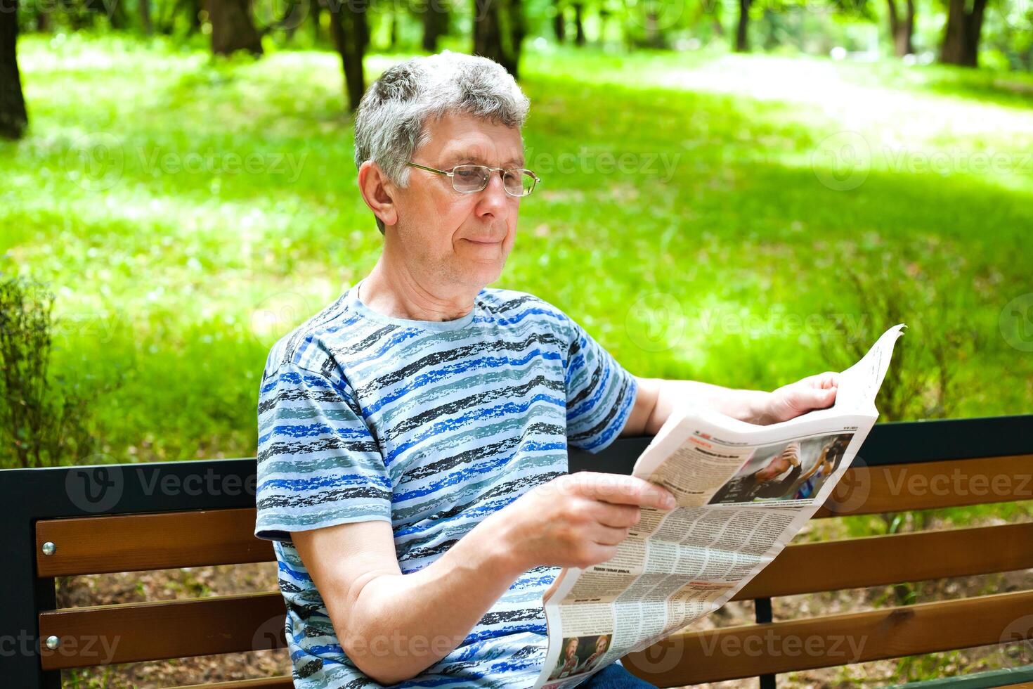 Portrait of senior man reading newspaper in the park on a bench. Summer sunny day. photo