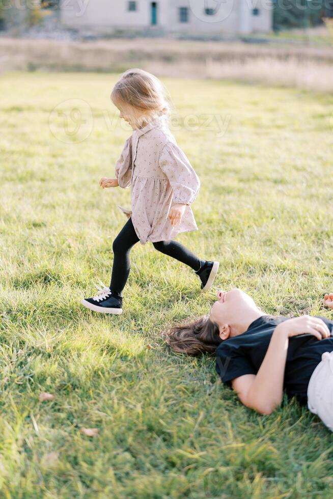 Smiling mother lies on the green lawn looking at a little girl running past photo