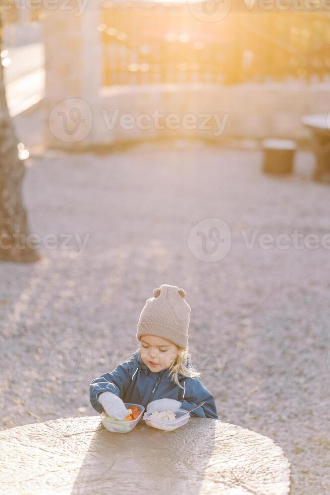 Little girl takes a piece of cucumber from a lunchbox while standing behind a large tree stump in the park photo