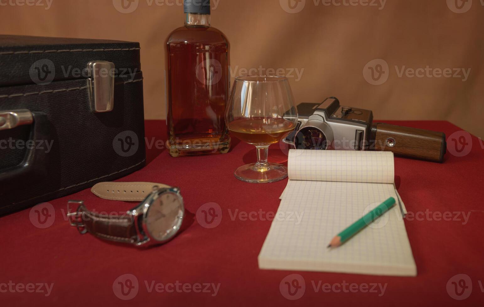Old camera, wrist watch, bottle, glass, notebook, pencil, suitcase. Red tablecloth, brown background - retro bystalation photo