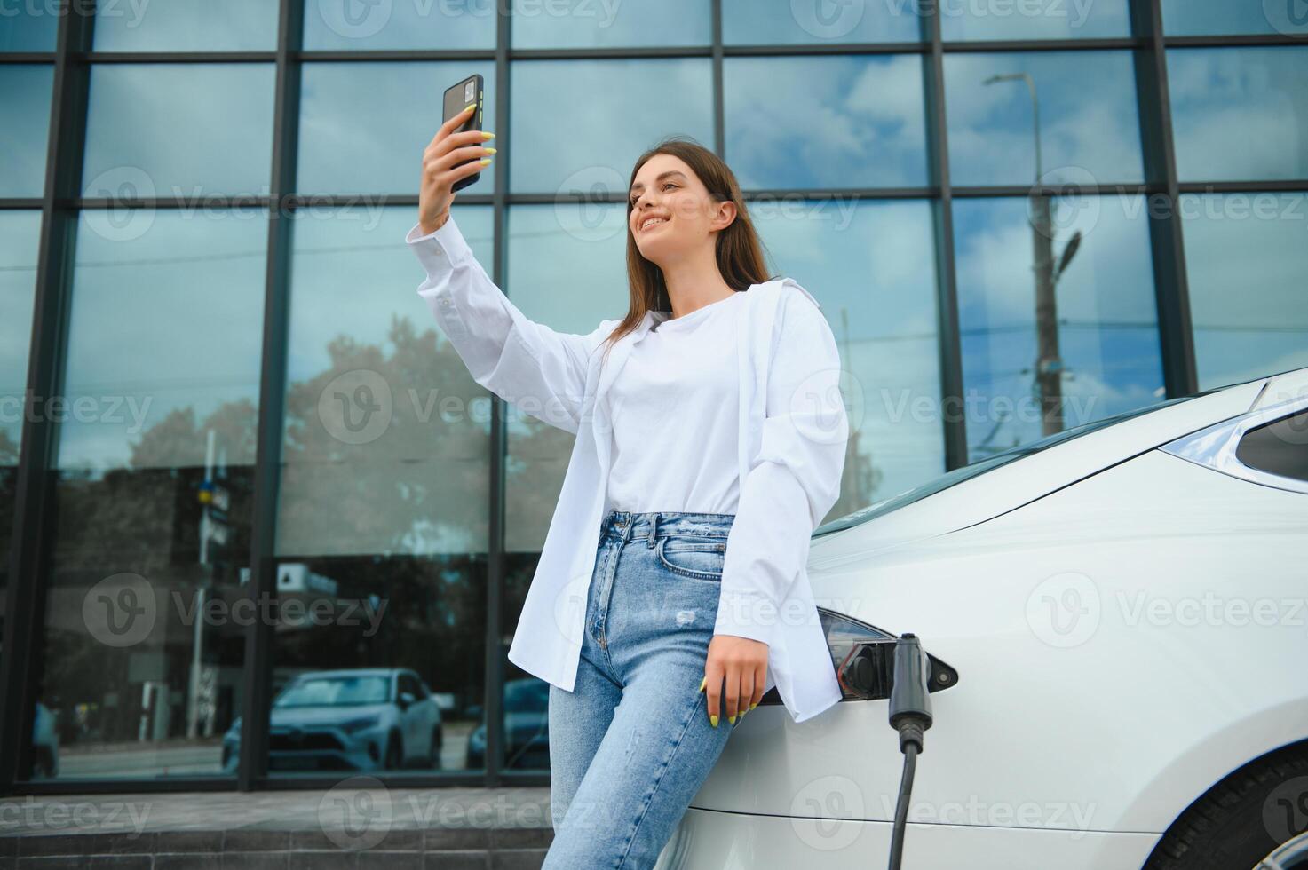 Phone in hands. Woman on the electric cars charge station at daytime. Brand new vehicle. photo