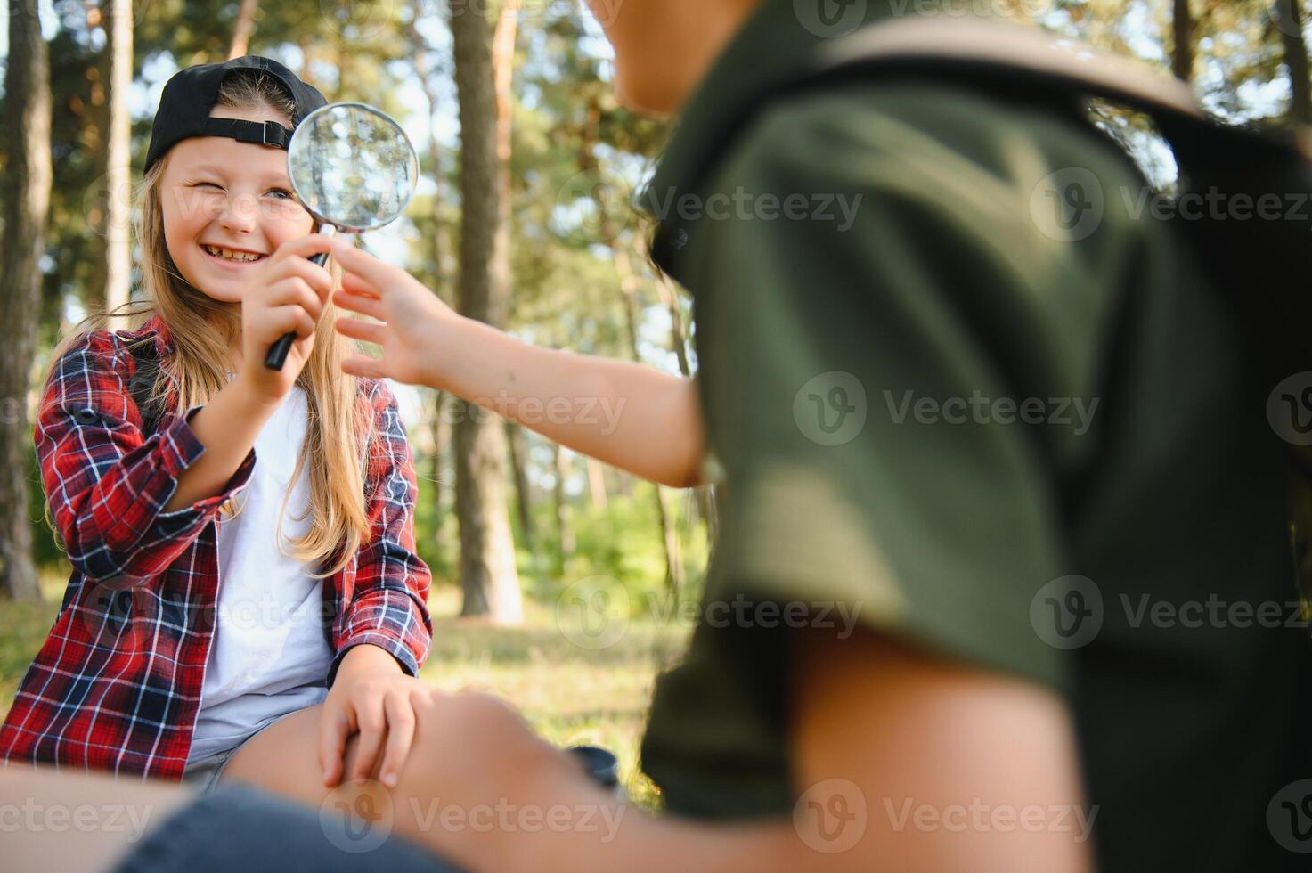 Group of curious happy school kids in casual clothes with backpacks exploring nature and forest together on sunny autumn day, girl holding magnifying glass and looking at fir cone in boy hands. photo