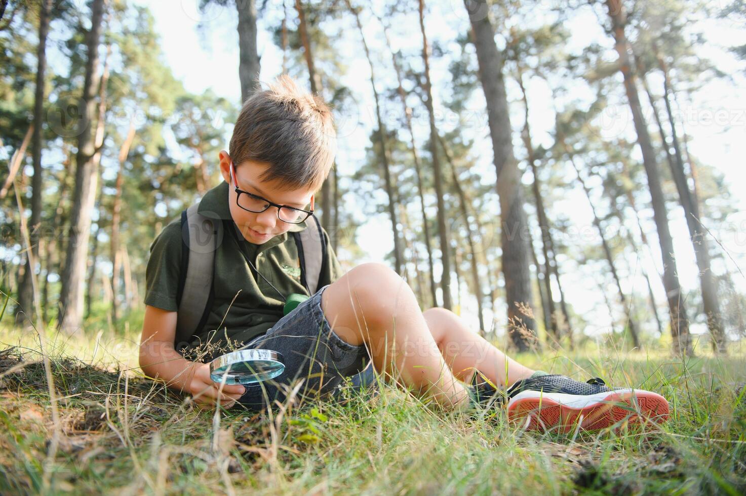 schoolboy is exploring nature with magnifying glass. Summer vacation for inquisitive kids in forest. Hiking. Boy-scout. photo