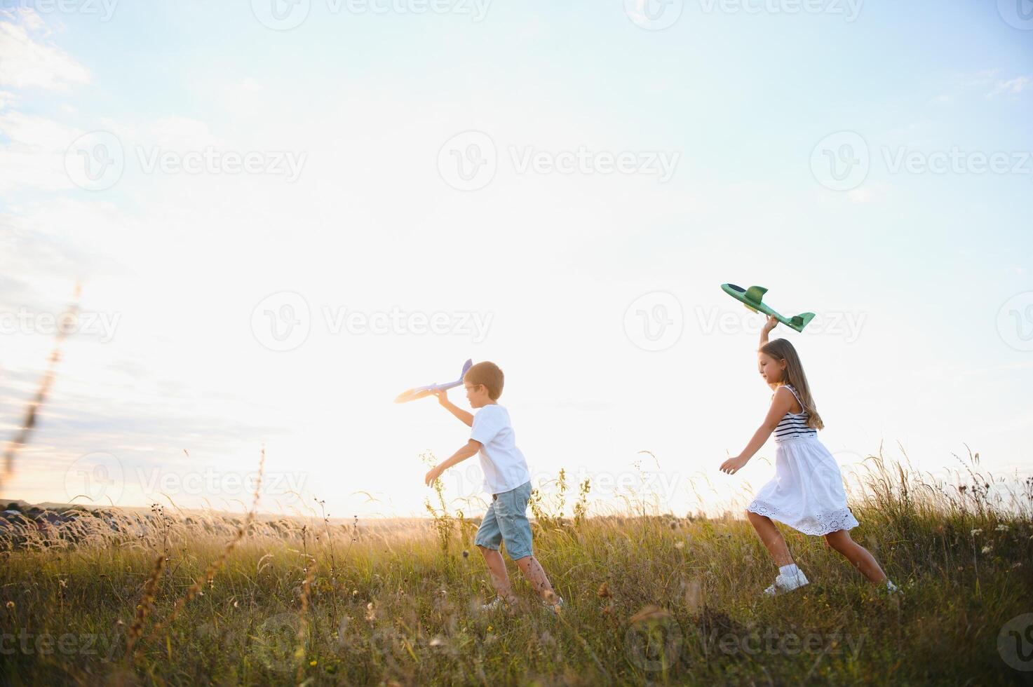 Running boy and girl holding two green and blue airplanes toy in the field during summer sunny day photo