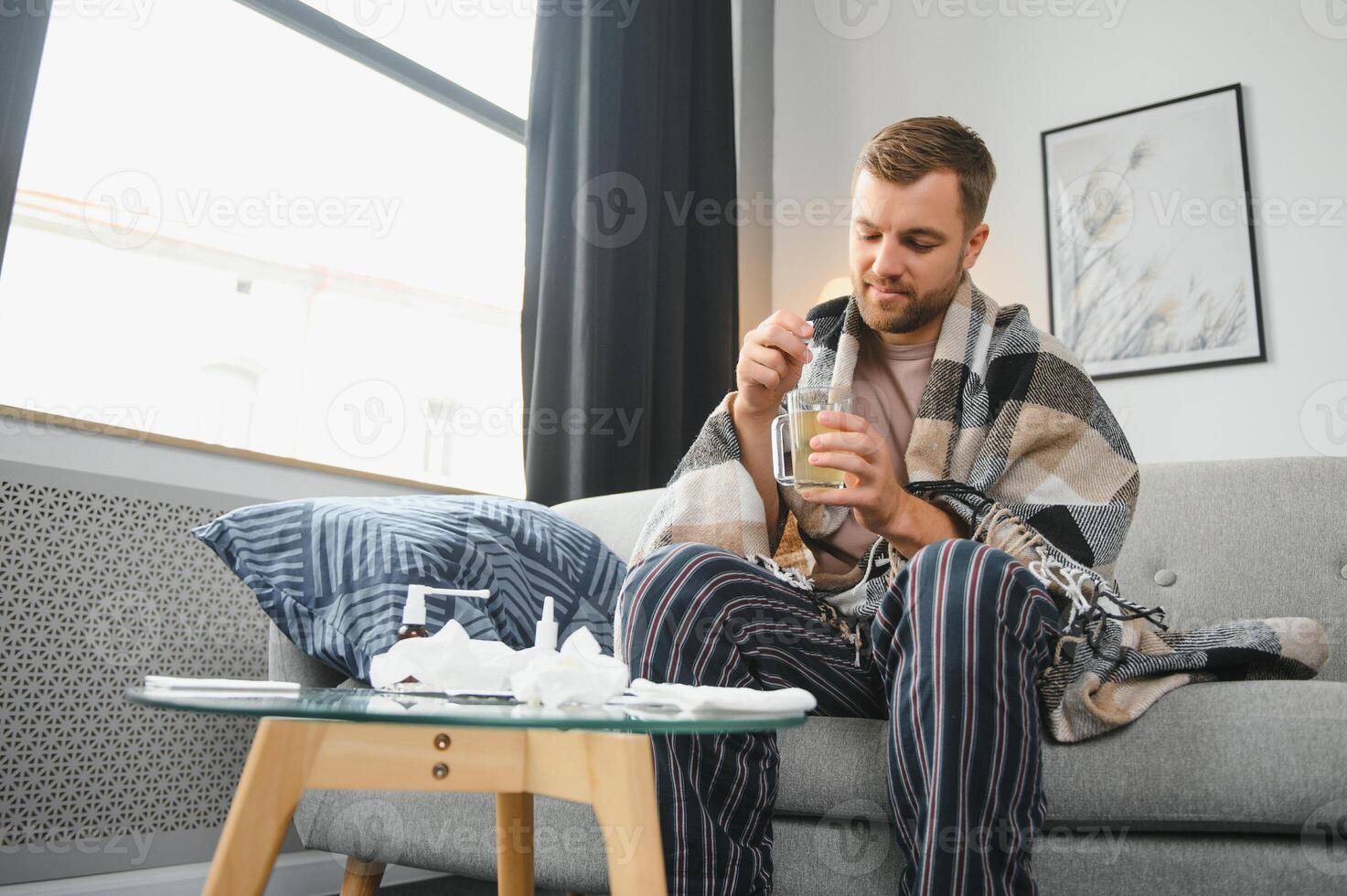Sick bearded man who has bad cold or seasonal flu sitting on couch at home. Guy with fever wearing warm plaid shivering with worried face expression. photo