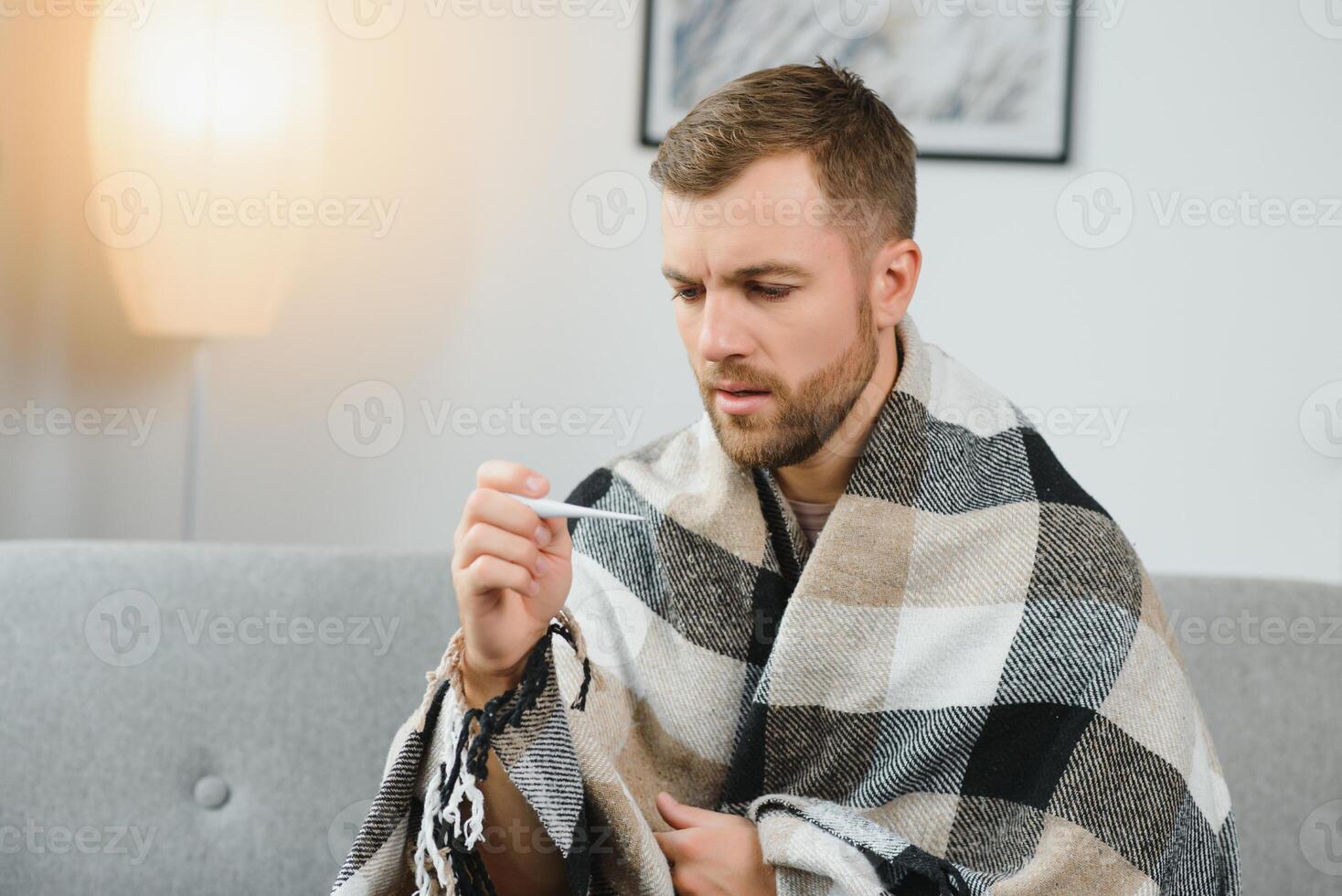 Sick adult man who has bad cold or seasonal flu virus is fighting illness at home. Unhappy middle aged man is sitting on sofa wrapped in warm scarf and plaid, holding paper tissue and sneezing. photo