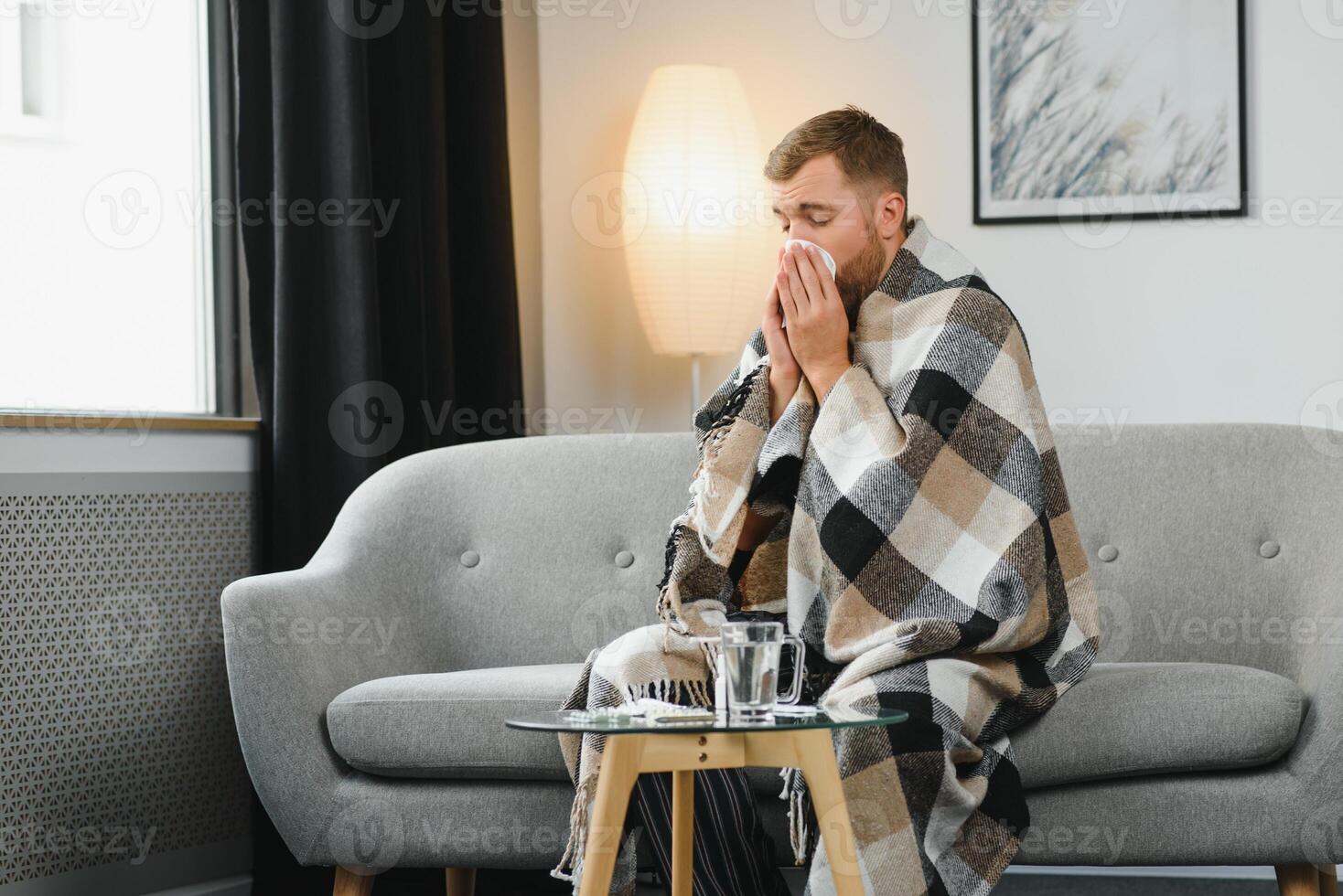 Sick adult man who has bad cold or seasonal flu virus is fighting illness at home. Unhappy middle aged man is sitting on sofa wrapped in warm scarf and plaid, holding paper tissue and sneezing. photo