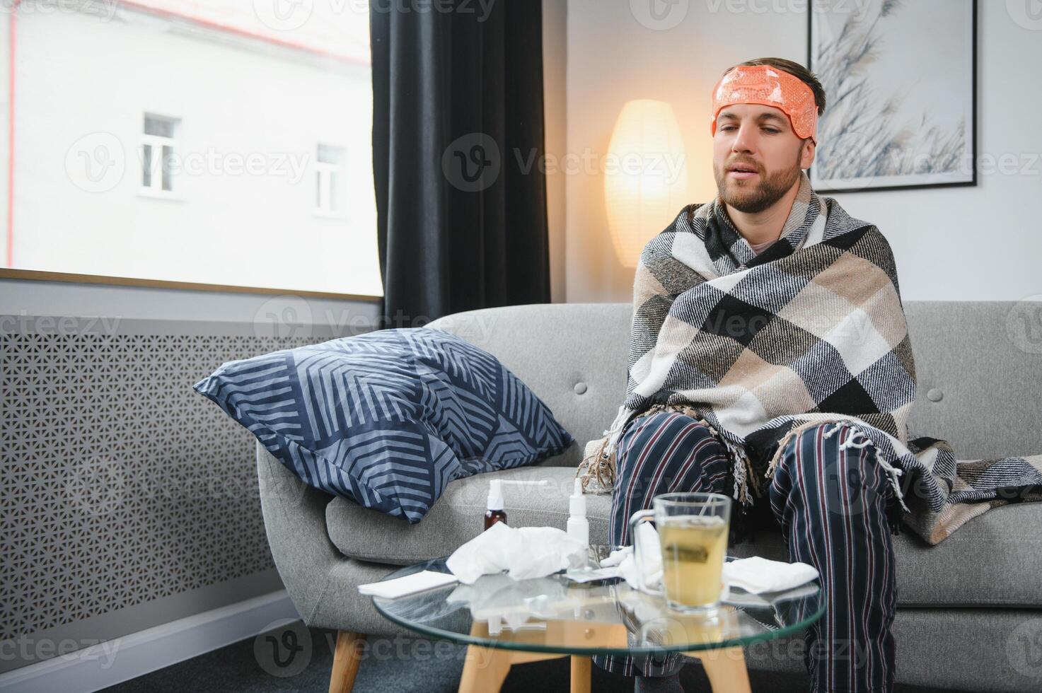 Sick bearded man who has bad cold or seasonal flu sitting on couch at home. Guy with fever wearing warm plaid shivering with worried face expression. photo