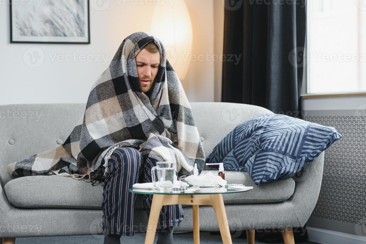 Sick adult man who has bad cold or seasonal flu virus is fighting illness at home. Unhappy middle aged man is sitting on sofa wrapped in warm scarf and plaid, holding paper tissue and sneezing. photo