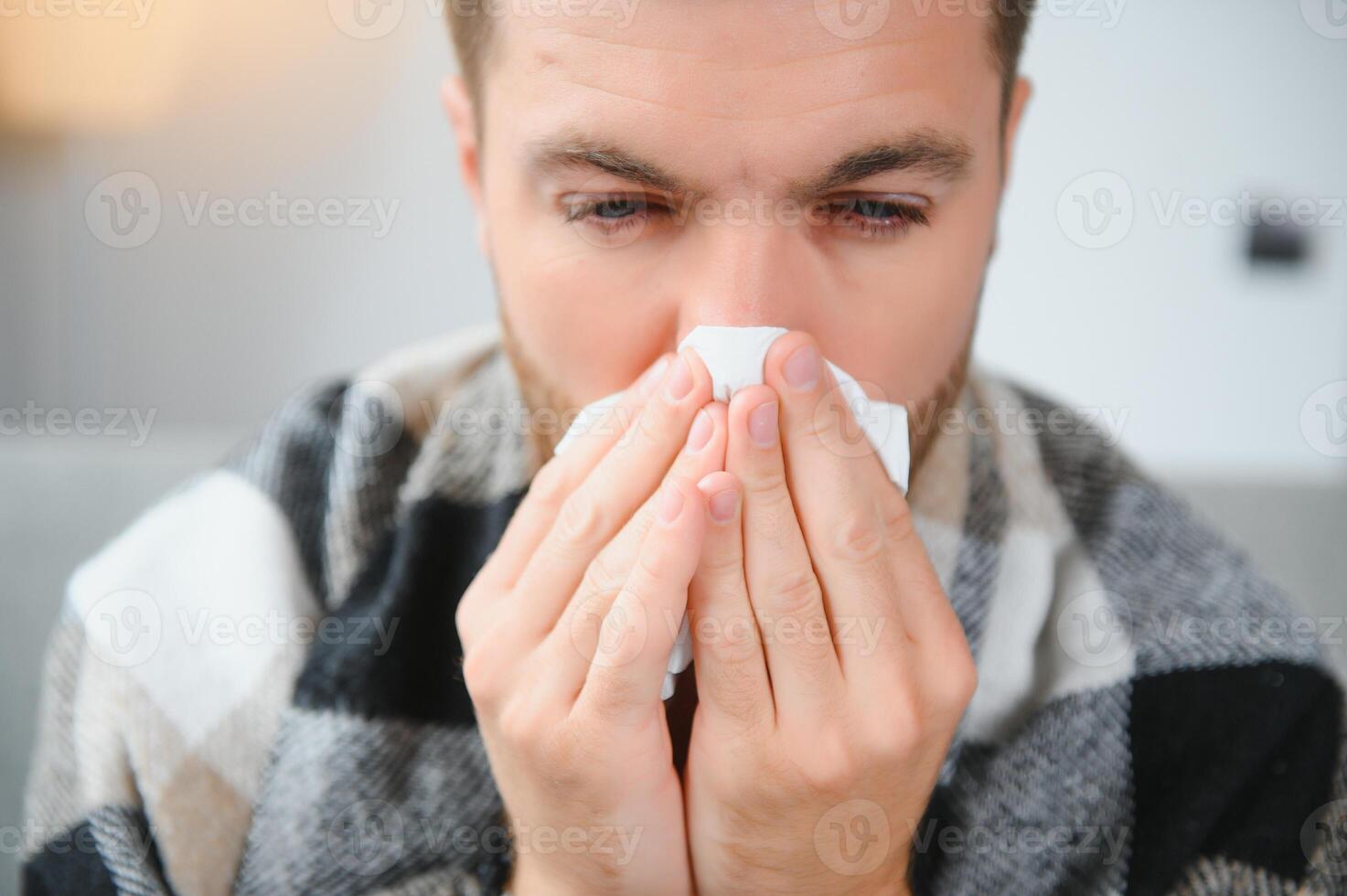 Young man suffering from a common cold and flu or allergy sit at home wrapped in blanket and wipe his nose with tissues while he have a strong headache pain, healthcare concept. photo