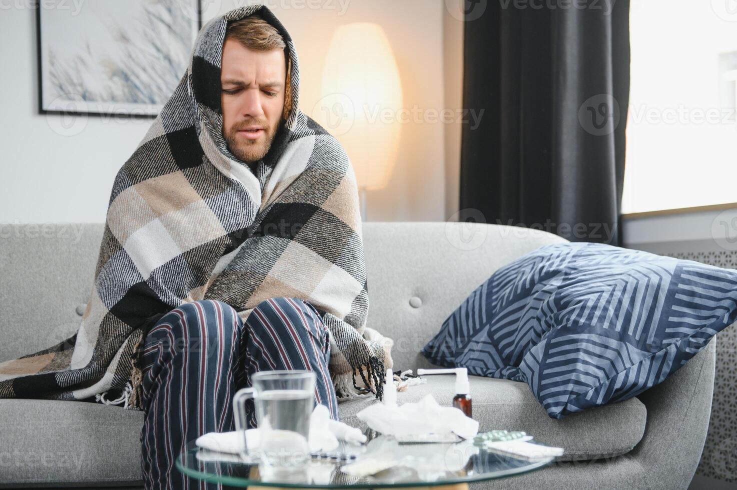 It's cold at home in wintertime. Man freezing in his house in winter because of broken thermostat. young guy wrapped in woolen plaid shivering while sitting on sofa in living room interior photo