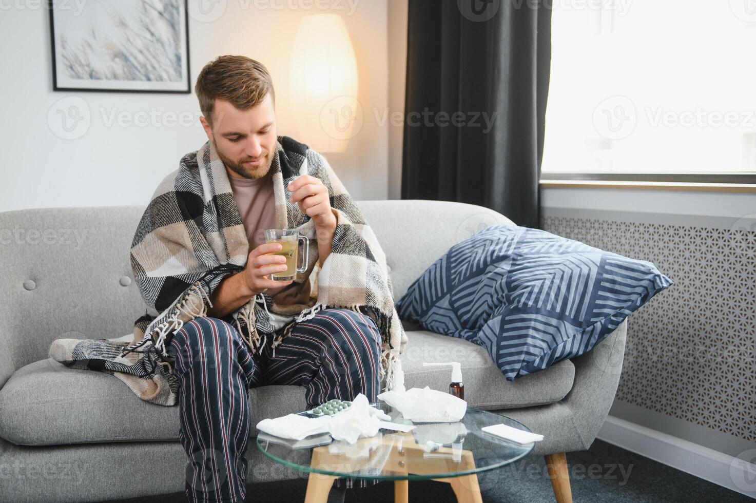 Sick bearded man who has bad cold or seasonal flu sitting on couch at home. Guy with fever wearing warm plaid shivering with worried face expression. photo