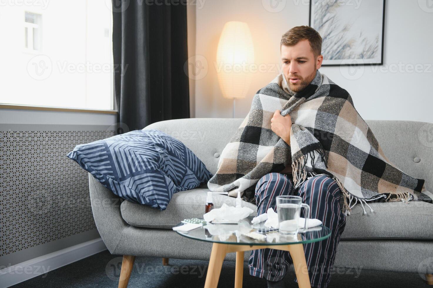 It's cold at home in wintertime. Man freezing in his house in winter because of broken thermostat. young guy wrapped in woolen plaid shivering while sitting on sofa in living room interior photo