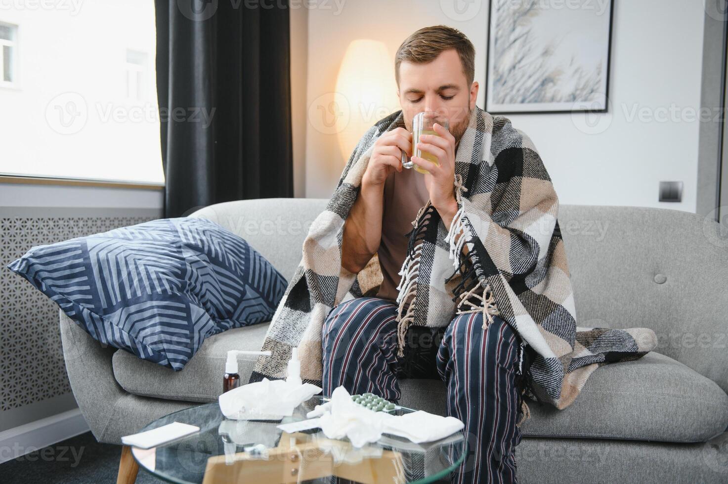 Sick bearded man who has bad cold or seasonal flu sitting on couch at home. Guy with fever wearing warm plaid shivering with worried face expression. photo