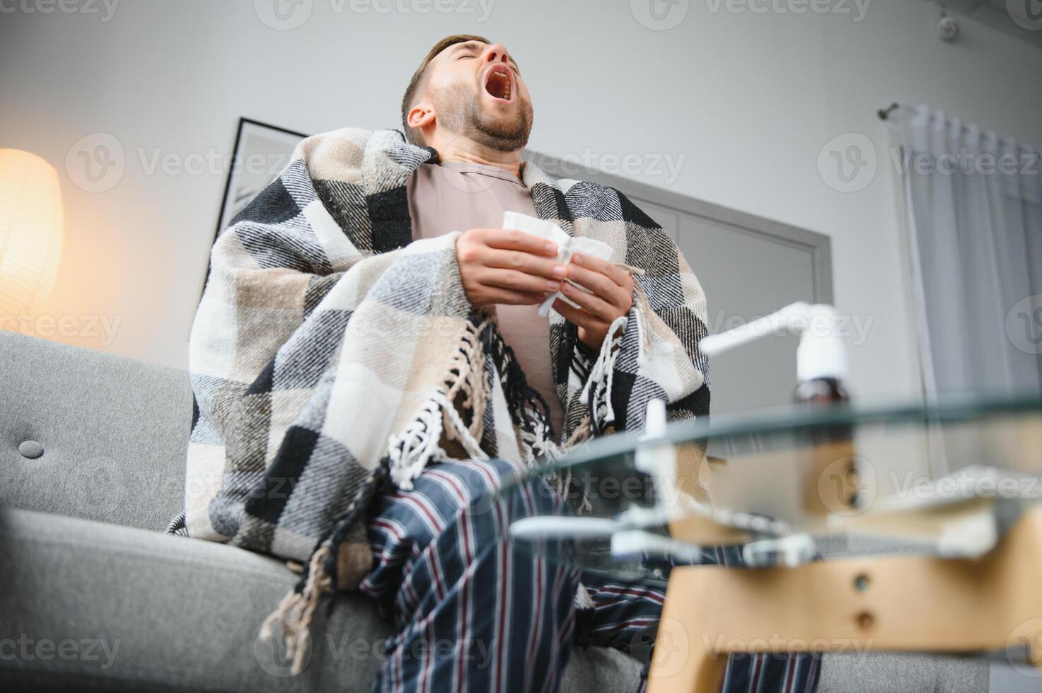 Young man suffering from a common cold and flu or allergy sit at home wrapped in blanket and wipe his nose with tissues while he have a strong headache pain, healthcare concept. photo