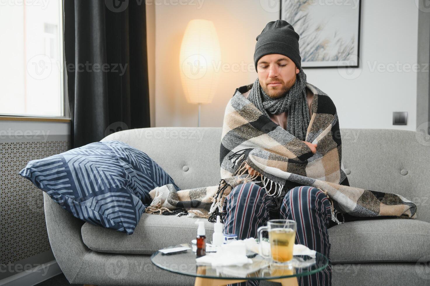 Sick bearded man who has bad cold or seasonal flu sitting on couch at home. Guy with fever wearing warm plaid shivering with worried face expression. photo