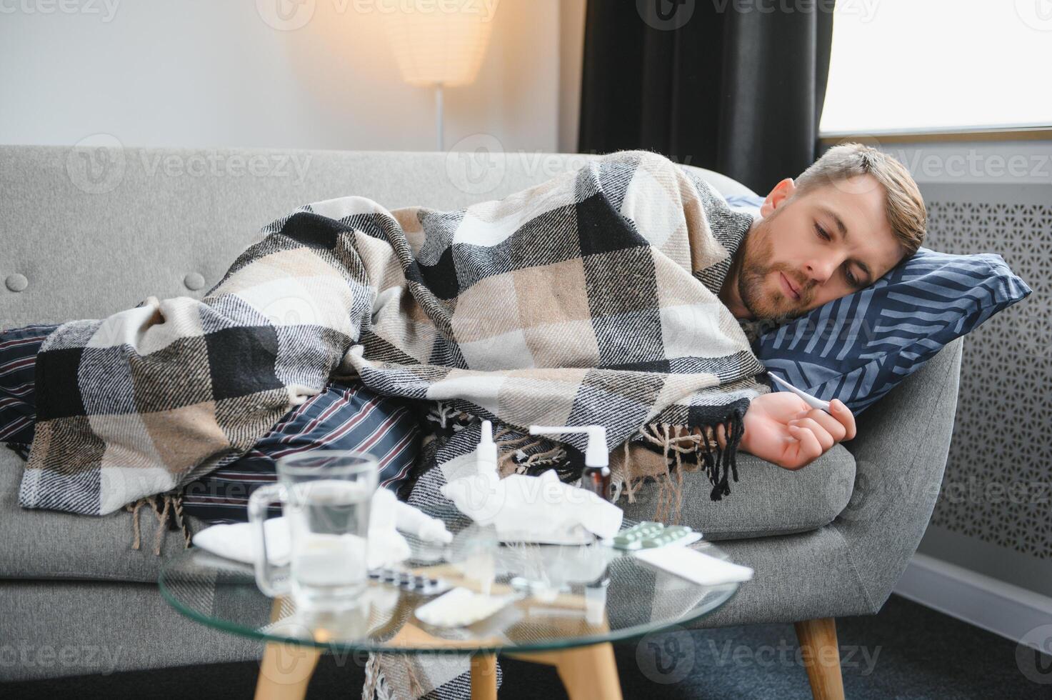 Sick bearded man who has bad cold or seasonal flu sitting on couch at home. Guy with fever wearing warm plaid shivering with worried face expression. photo