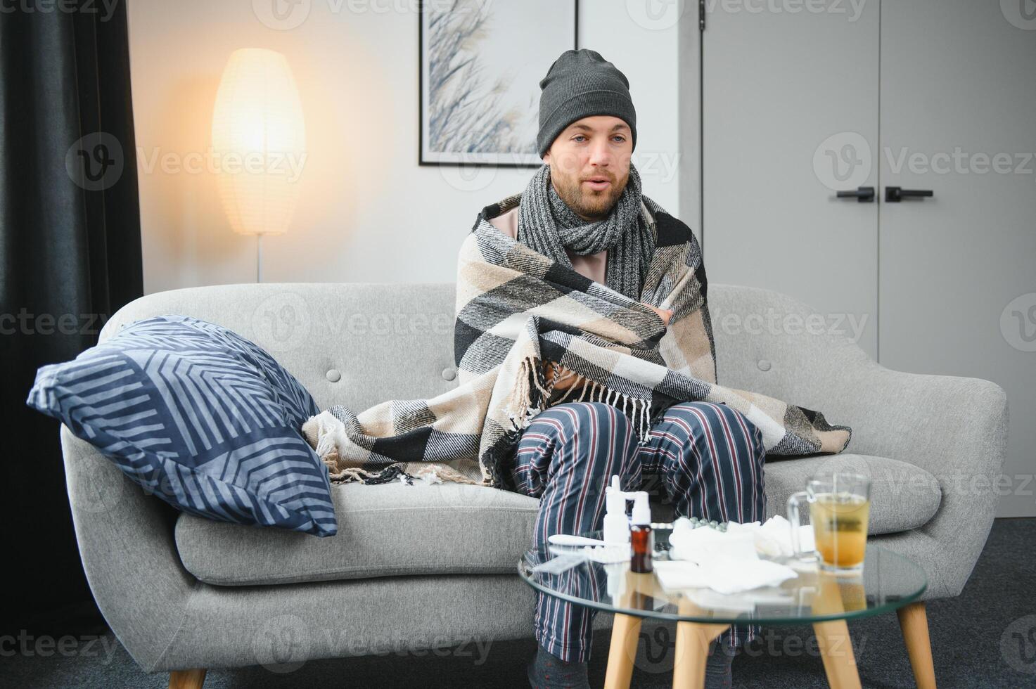 It's cold at home in wintertime. Man freezing in his house in winter because of broken thermostat. young guy wrapped in woolen plaid shivering while sitting on sofa in living room interior photo