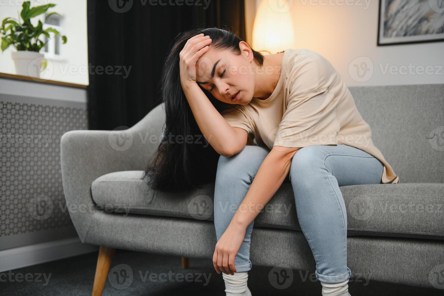 Portrait of beautiful young woman with depressed facial expression sitting on grey textile couch. Cyber bullying victim concept. Sad female in her room. Background, copy space photo