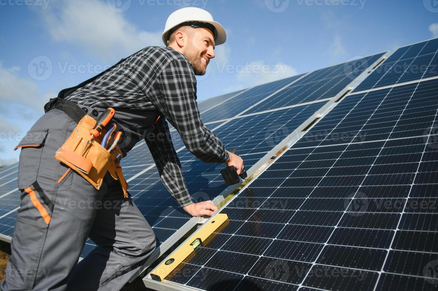 Portrait of smiling confident engineer technician with electrical screwdriver, standing in front of unfinished high exterior solar panel photo voltaic system
