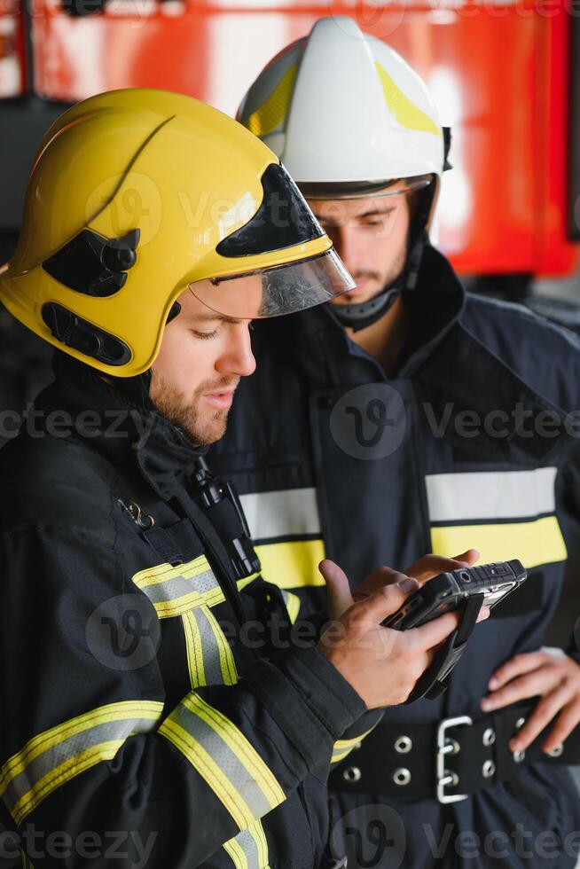 Two professional firefighters with uniforms and protective helmets posing infront of a firetruck. photo