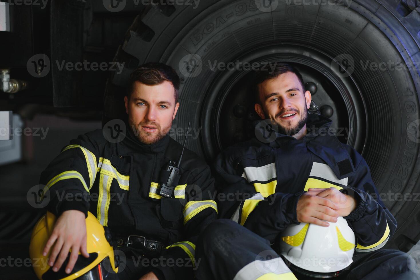 Two professional firefighters with uniforms and protective helmets posing infront of a firetruck. photo