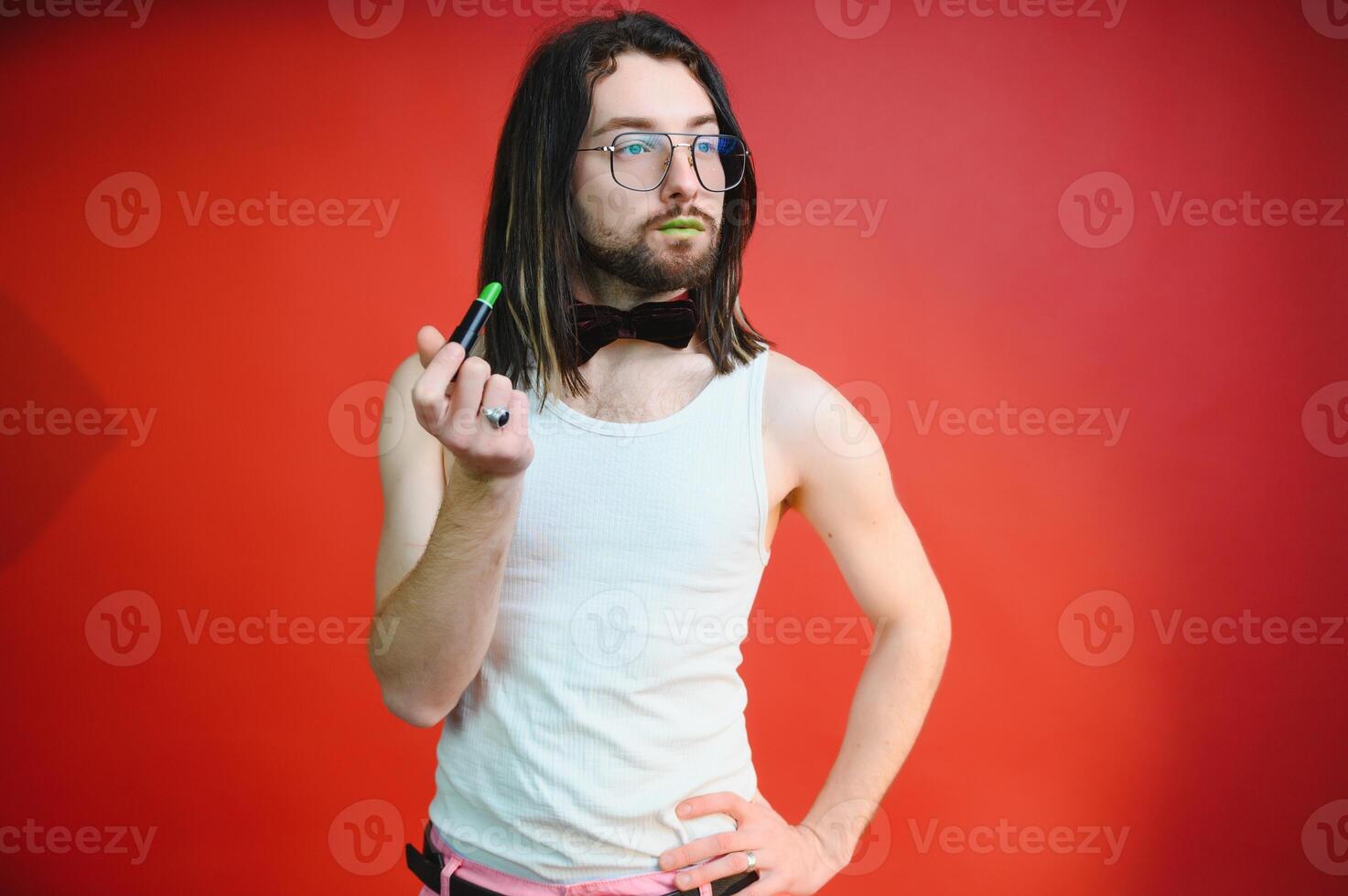 Young man applying lip gloss on his lips and looking in camera like mirror. Gay man doing make up against red background photo