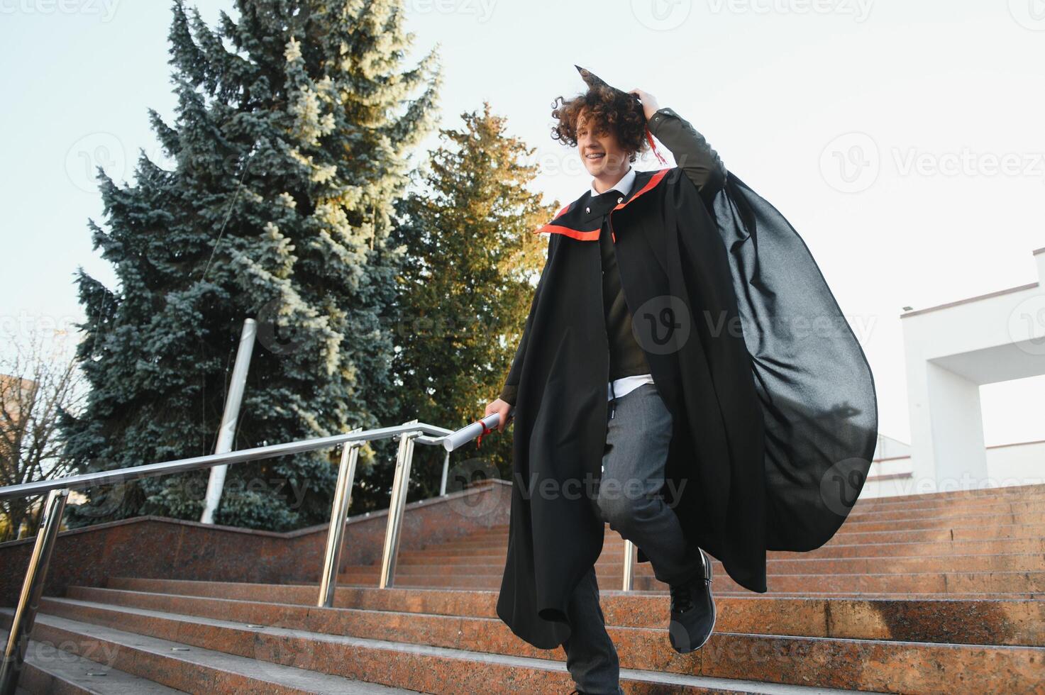 Happy smiling university graduate in mantle holding diploma in raised hand and expressing happiness over university building at background. Successful graduating from university or college photo