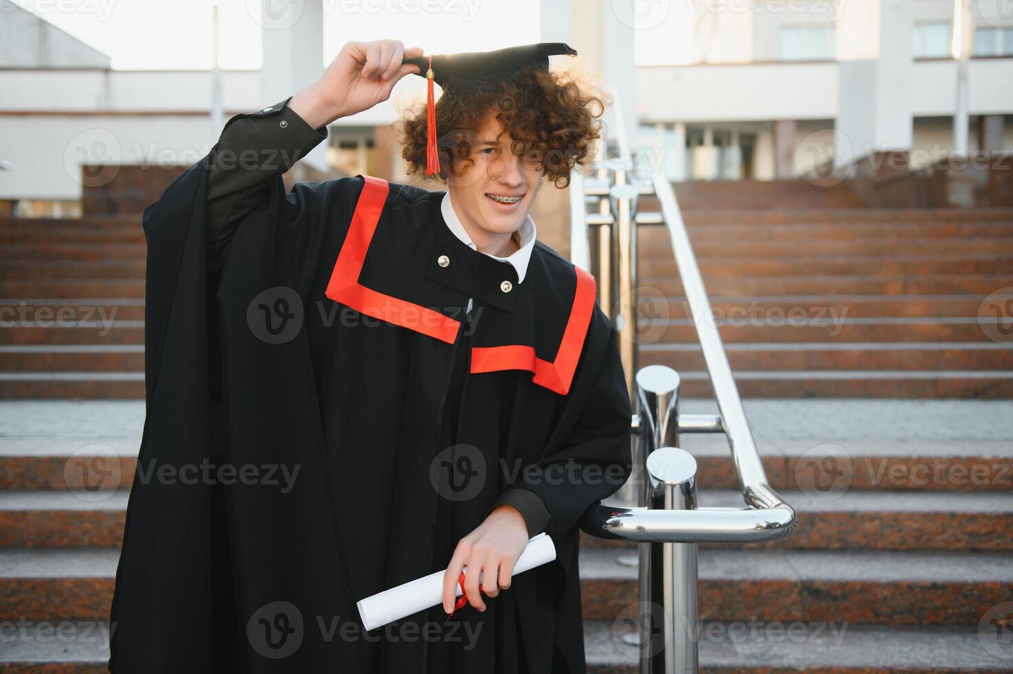 Graduation from university. Young smiling boy university graduate in traditional bonet and mantle standing and holding diploma in hand over university building background photo