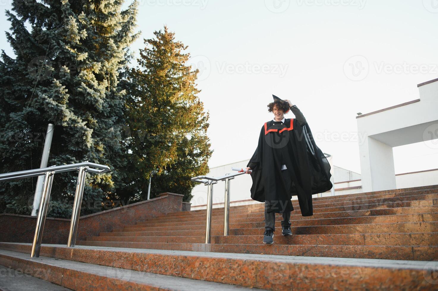 Graduation from university. Young smiling boy university graduate in traditional bonet and mantle standing and holding diploma in hand over university building background photo