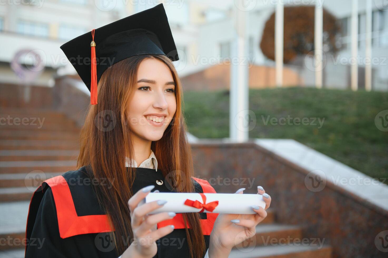 Happy cute brunette caucasian grad girl is smiling. She is in a black mortar board, with red tassel, in gown, with nice brown curly hair, diploma in hand. photo