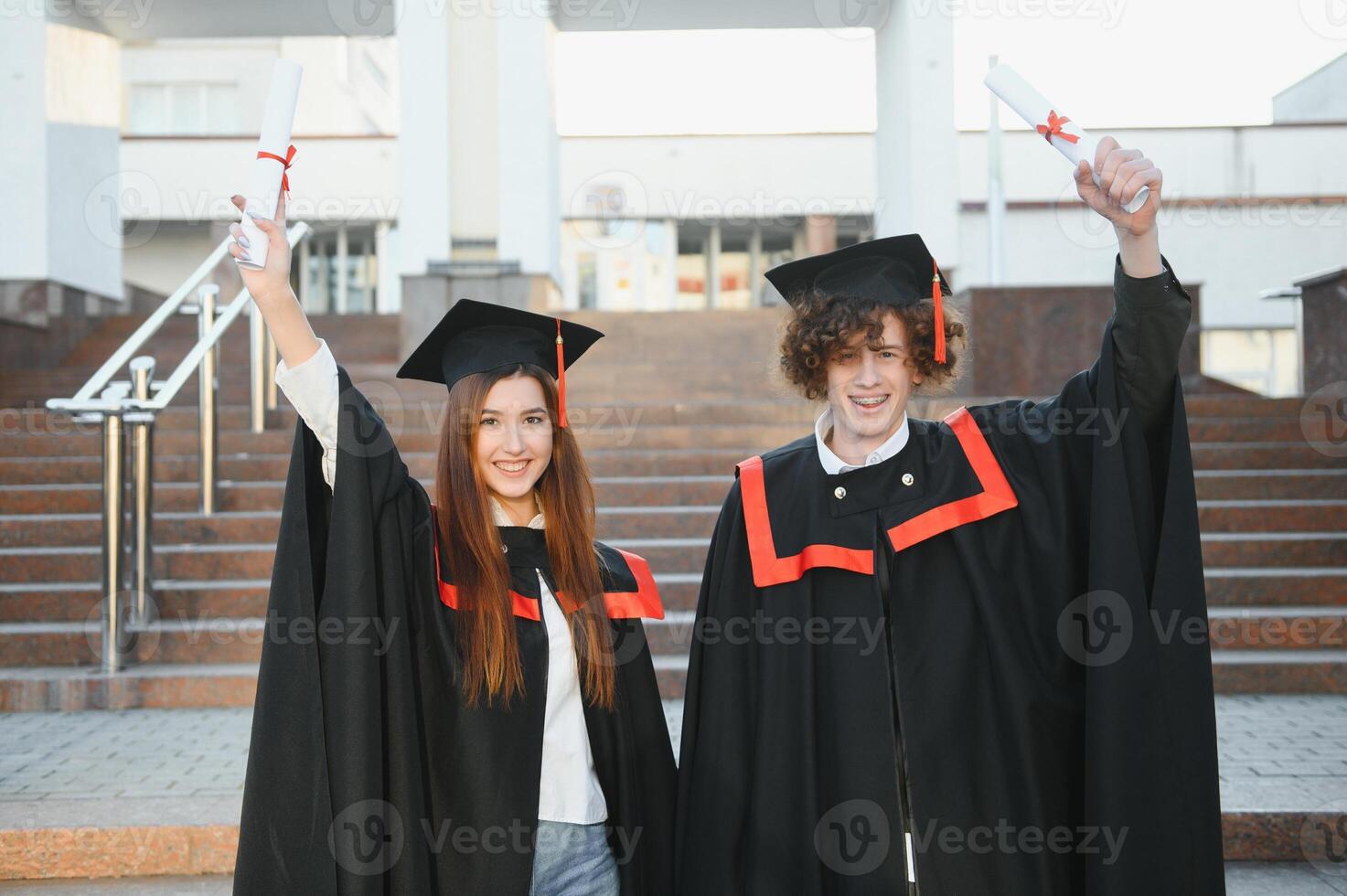 Portrait of happy graduates. Two friends in graduation caps and gowns standing outside university building with other students in background, holding diploma scrolls, and smiling photo