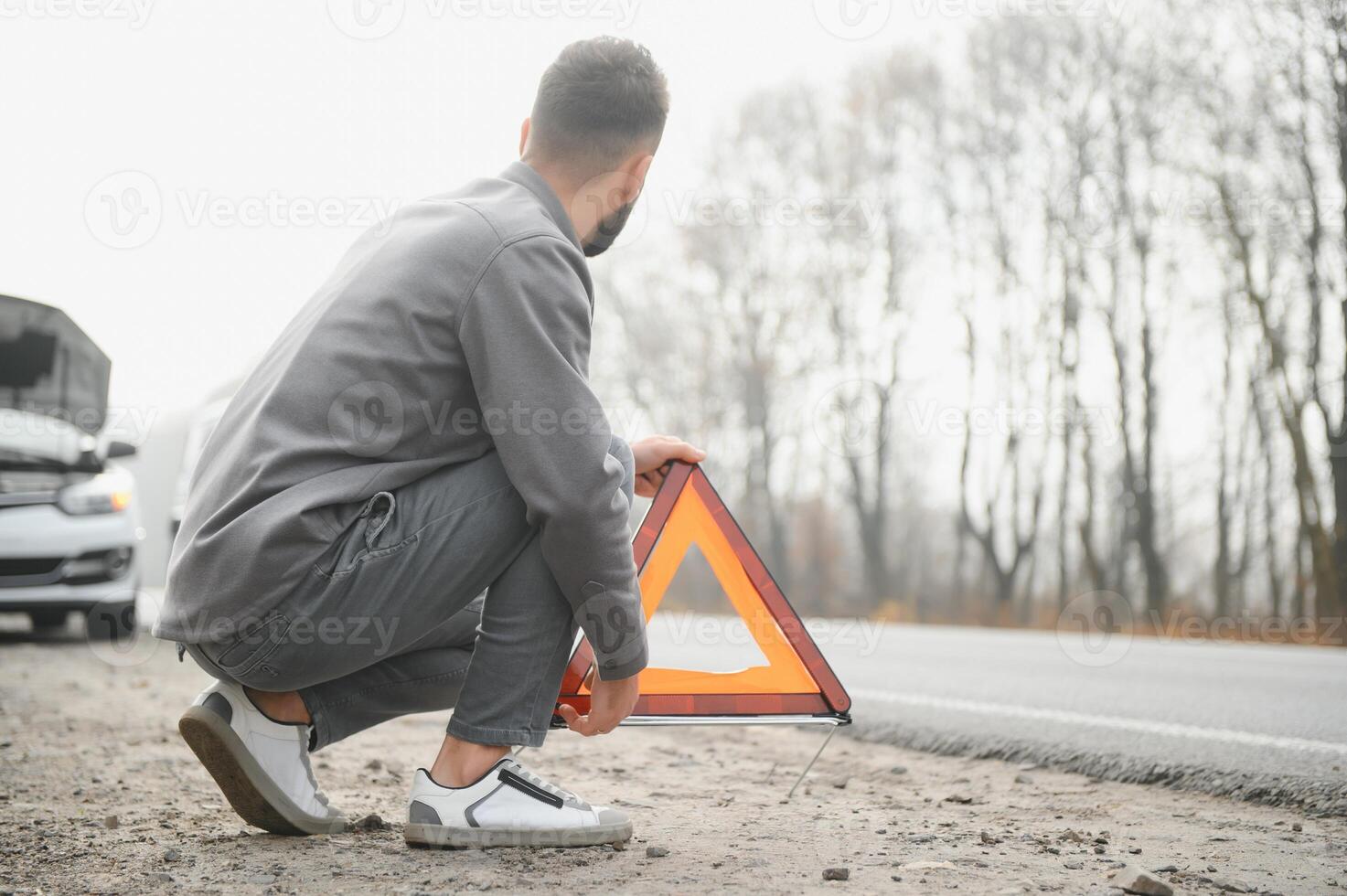 A young man near a broken car with an open hood on the roadside. photo