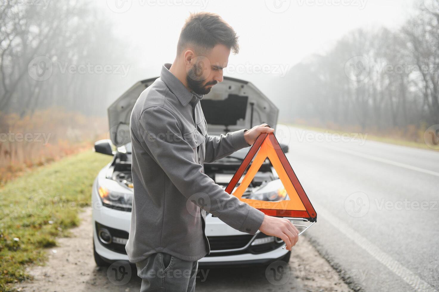 A young man near a broken car with an open hood on the roadside. photo