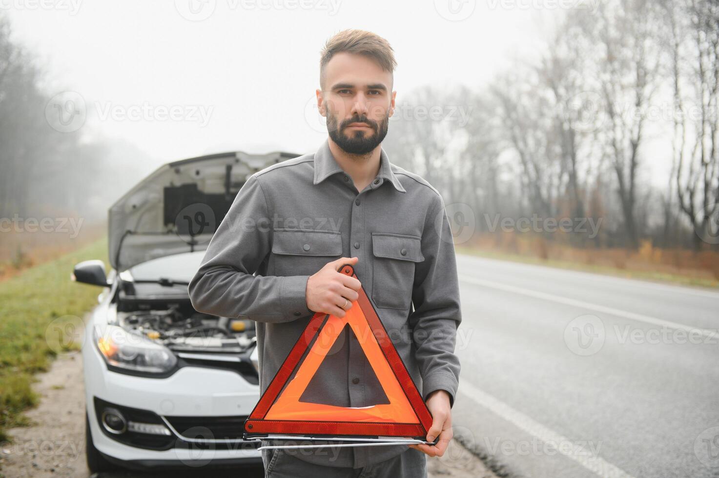 A young man near a broken car with an open hood on the roadside. photo